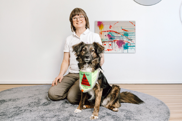 Eine Frau mit Brille kniet auf einem grauen Teppich neben einem braun-schwarzen Hund in einer grünen Weste. An der weißen Wand hinter ihnen hängt ein farbenfrohes abstraktes Gemälde.