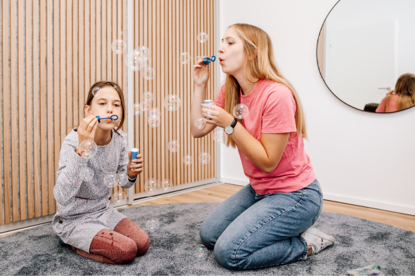 Zwei Mädchen knien auf einem grauen Teppich in einem modernen Zimmer in Kiel und lassen gemeinsam Seifenblasen steigen.
