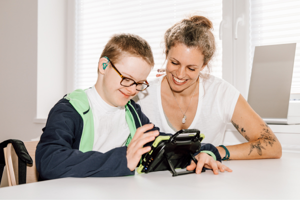 Ein kleiner Junge mit Brille und Hörgerät bedient an einem Tisch ein Tablet, während eine Frau ihm aufmunternd zulächelt. Sie befinden sich in einem Raum in Kiel.